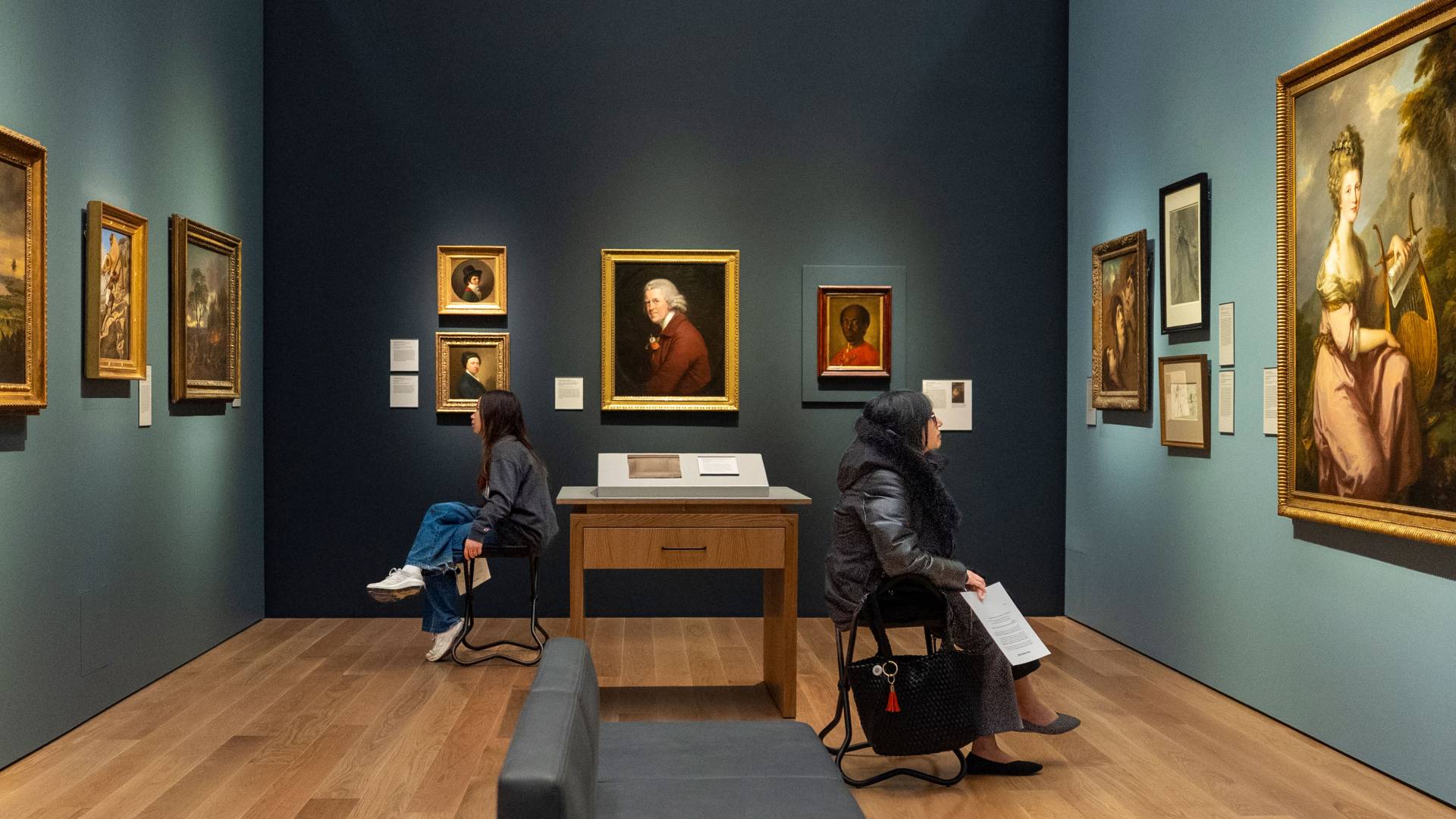 Two people sit and look closely at paintings on the wall